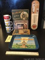 Full view of vintage advertising decor items on table including Land O'Lakes tray, thermometer, H.J. Heinz wall plaque, mugs, Carnation tins, Jello box, and NE Blocks. Ruler for scale.
