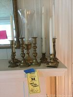Seven brass candlesticks displayed on a white shelf including styles with tapered candles and glass hurricane covers, showing a variety of base shapes and sizes.