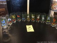 Full set of 12 Anchor Hocking 12 Days of Christmas glasses arranged in a row on black surface with measuring ruler behind for scale. Each glass has colorful day-specific Christmas illustrations.