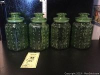 Four green glass canister jars on black surface, showing the embossed scroll pattern and sealed lids.