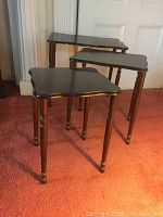 Set of 3 nesting tables arranged largest to smallest showing wood legs and dark brown composition tops with scalloped edges and gold trim.