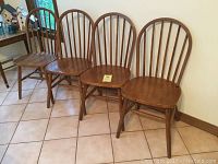 Four wooden Windsor style dining chairs with bow back design, viewed side by side against a wall on tile floor.