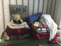 Stack of assorted throws and comforters on green bench with background wallpaper.