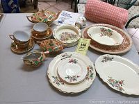 Full overview of vintage china collection showing lusterware tea set, floral plates, Bunnykins bowl, Syracuse platter, Beech Nut sticker book and tablecloth rolled up.