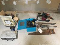 Wide shot of assorted workshop and garden hand tools, power tools and storage containers grouped on the floor.