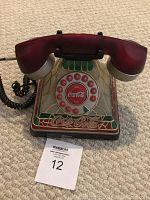 Front view of Coca Cola decorative rotary dial telephone with handset on carpet. Shows stained glass-style design and Coca Cola logos on dial and base.