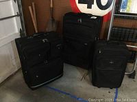 Three black fabric suitcases of varying sizes, all upright with visible handles and wheels, positioned on a floor with various other items in background.