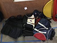 Overall view showing assortment of bags including Samsonite black garment bag, white handbag, navy blue rolling bag, and other backpacks on floor against pegboard wall.