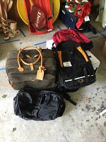Group photo showing all items: brown BRICS leather carry-on suitcase, matching black computer bag, black daypack with orange detail, red backpack, small black duffel.