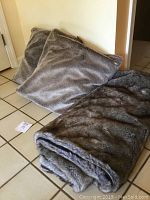 Two faux lynx pillows and a matching blanket arranged on a tiled floor, showing their natural fur texture and color.