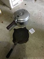 Photo showing Presto Cook Master aluminum stovetop pressure cooker with black handle and cast iron Jotul waffle iron with handle on concrete floor.