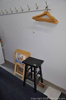 Wide view showing black wooden stool next to two stacked folded TV trays and six wood hangers hanging on large wall hooks.