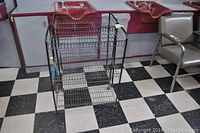 Black wire basket cart on casters shown from front, placed on checkered floor with surrounding furniture.