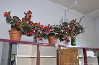 Wide view showing three terra cotta pots with faux floral arrangements and one green glass vase on a high shelf.
