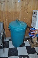 Photo of large teal Rubbermaid trash bin with lid and clamping handles, standing on checkered floor.