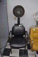 Front view of black cushioned chair with attached hooded hair dryer for drying hair, standing on metal frame with armrests against a wall with power outlet.