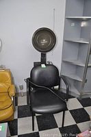Black salon chair with attached hooded hair dryer station, placed indoors on checkered floor next to shelves and a mustard-colored chair.