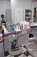 Small office chair with black hair dryer attachment on the seat in front of salon counter holding salon supplies including Barbicide jars, hair sprays, and curling irons
