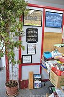 Photo showing framed signs including a black barbershop price list, two framed motivational quotes, barbershop birdhouse wall decor, and framed group photographs in black and dark wood frames on a white and red wall.