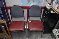 Two metal frame chairs with burgundy vinyl seats and dark gray cloth backrests placed next to each other in front of a red wall-mounted shelf and a world map.