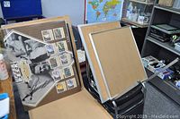 Two cork boards with silver metal frames leaning against stacked chairs, with an empty silver frame and several posters including a black and white hairstyle salon poster.