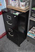 Side angled photo of the black metal two drawer filing cabinet showing front and one side view with details of handles, lock and wear marks.