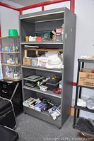 Tall gray metal storage cabinet with 4 dropleaf doors and open shelves containing various small items, shown with wear and marks from use. Cabinet has side coat hook and locking mechanism without key.