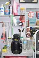 Overall view showing Bosch coffee machine on counter with other coffee station supplies, including packaged cleaning sponges, disposable coffee cups on a metal stand, water bottles, and cleaning sprays on shelf above.