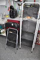 Photo showing two folding step ladders including one with a paint tray, small foldable step stool, and three stacked black folding chairs in a workroom setting.