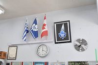 Wall display showing three small flags on stands (Greek, Toronto Maple Leafs, Canadian), a framed Toronto Maple Leafs sign, and two wall clocks including a quartz clock and a saw blade clock.