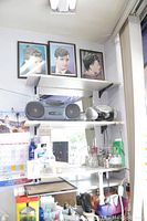 View showing Venturer cassette tape player, Electrohome CD player on shelves along with various barber supplies including combs and clips on surface below and three framed barber portraits above.