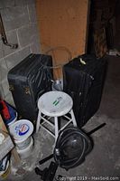 Photo of two black vintage suitcases, round white-painted wooden stool, black vacuum cleaner with hose and attachments, and two metal tomato cages in a basement or storage area.