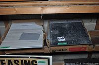 Two boxes on a wooden shelf, containing linoleum tiles in light gray/beige and black/charcoal colors.