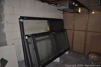 Overall view of the exterior shop window panel leaning against a basement wall, showing the black metal frame and glass top portion.