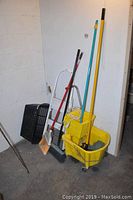 Wide view of four cleaning tools leaning against a wall: two brooms, a mop in a yellow mop bucket with wringer, a white folding step stool, and the black laundry basket placed on the ground to the side.
