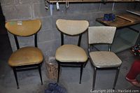 Three vintage chairs with metal frames and vinyl seats of different shades placed against a concrete wall. Visible wear and damage on the vinyl surfaces.
