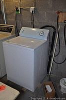 Full view of the white Kenmore washing machine positioned in a basement with cinder block walls, showing the appliance and surroundings such as utility hookups and another laundry machine beside it.
