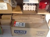 Boxes of white thermal printer tape rolls stacked in cardboard compartments with stacks of flat cardboard pieces on top of a large Uline Clear Shopper carton box.