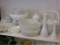 Shelf display showing multiple milk glass bowls, vases, hobnail bowl, lidded dish, and handled basket in white opaque glass.