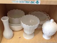 Milk glass bowls, dishes, and vases grouped on a shelf showing variety of shapes and sizes with scalloped edges and textured patterns.