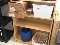 Shelving unit showing three shelves with various items placed on and below them (items not included). The unit is light woodgrain in color and appears to be in good condition.