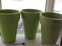 Three green plastic planters aligned on a white windowsill, lit by natural light from behind. All three planters have a tapered shape with a wider top opening and an outward lip rim. A power cord is seen lying in front of the planters.