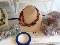 Photo of red multi-strand glass bead necklaces on display bust with sealed bags of green glass necklaces on the sides, blue painter's tape roll on table