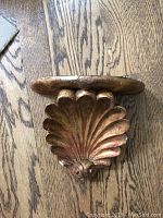 Close-up of the wall shelf showing the golden shell-shaped base and the wooden ledge top on a wood floor surface.