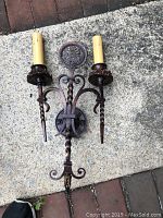 Photo of a dual-arm old world wrought iron style wall light with candle-shaped light holders and ornate scroll and floral iron details.