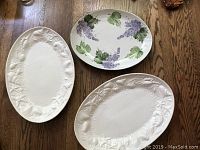 Three large serving platters on wooden floor: one grape-decorated plate and two white embossed platters with marine life themes.