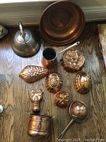Copper kitchenware and decorative molds laid out on wooden floor including kettle, bowl, ladle, cylindrical container, and nine decorative copper molds.