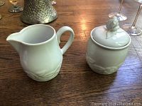 Cream and sugar set displayed on wooden surface, showing cream pitcher and sugar bowl with lid featuring bird knob.