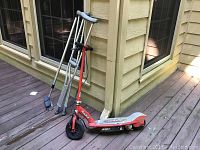 Photo of red Razor electric scooter alongside metal crutches and orange-handled extendible broom on porch deck.
