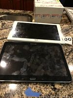 Two tablets on countertop, one white Acer and one black Samsung, showing screen and size comparison.